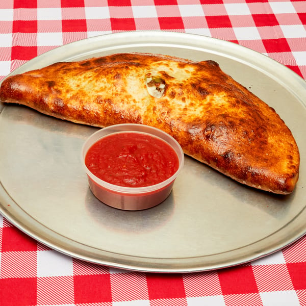 Golden-brown calzone with a side of red marinara sauce on a metal tray over a red and white checkered tablecloth.