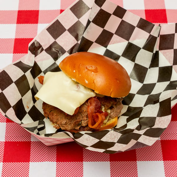 A cheeseburger with bacon on a brioche bun in a checkered paper tray on a red and white tablecloth.