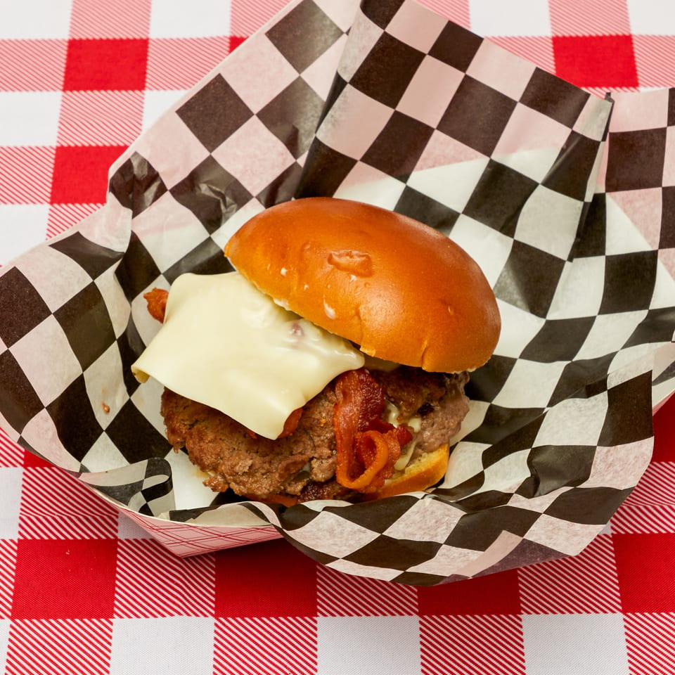A cheeseburger with bacon on a brioche bun in a checkered paper tray on a red and white tablecloth.