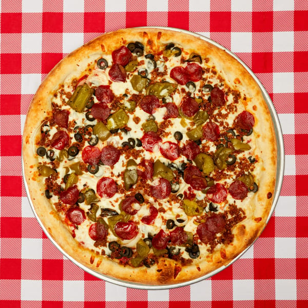 A whole pizza with pepperoni, green peppers, black olives, and ground beef on a red and white checkered tablecloth.